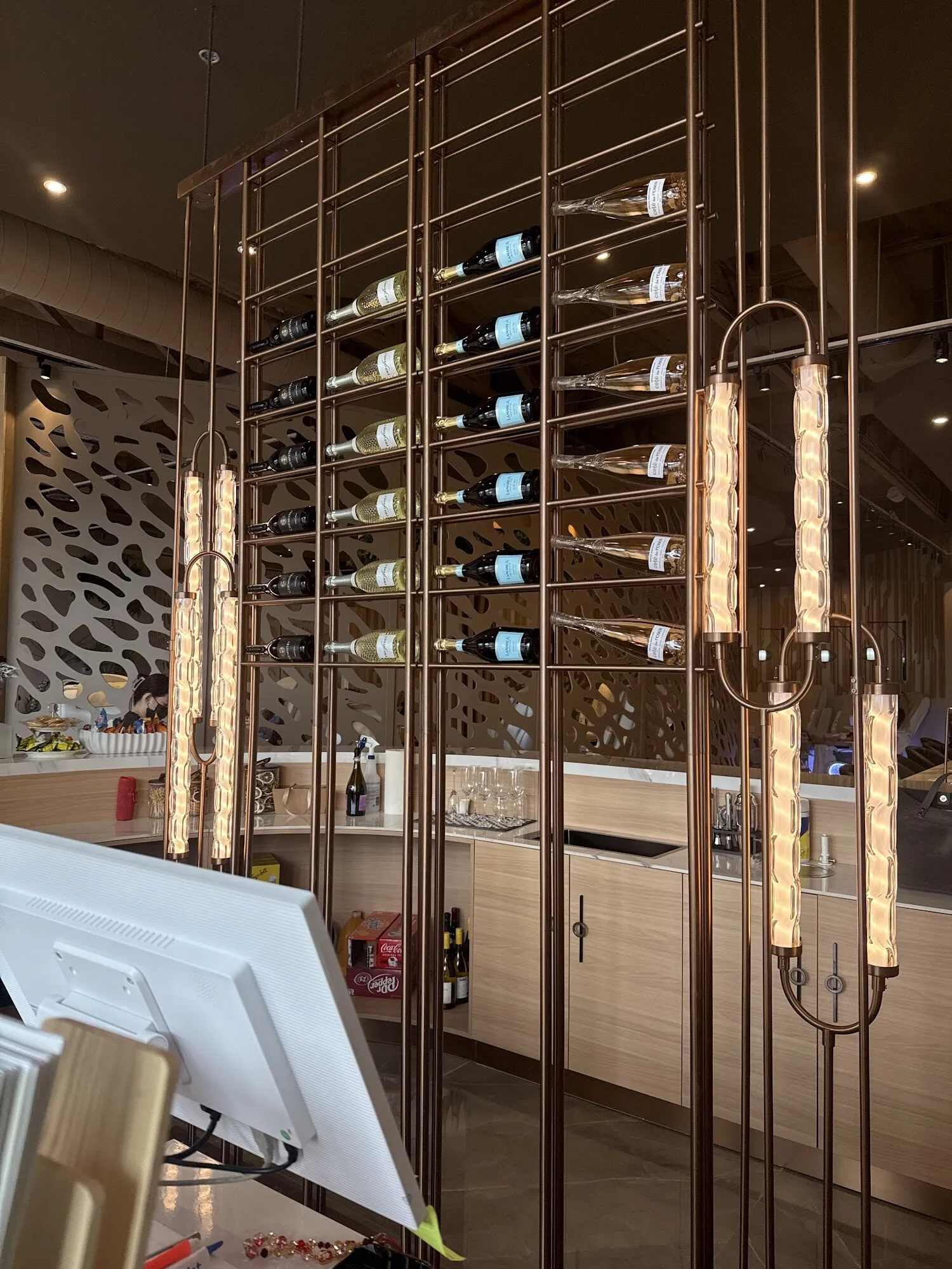 A bronze rack of bottles lit by tall pendant lights beside the bar at Zara Nail Bar Buckeye.