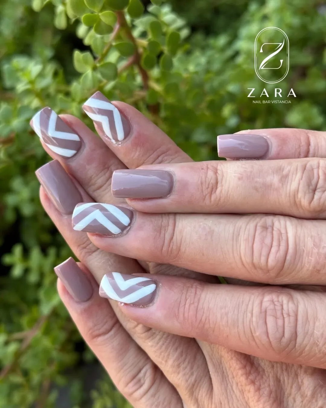 Short square nails in a mauve taupe with white chevron accents, photographed against green foliage.