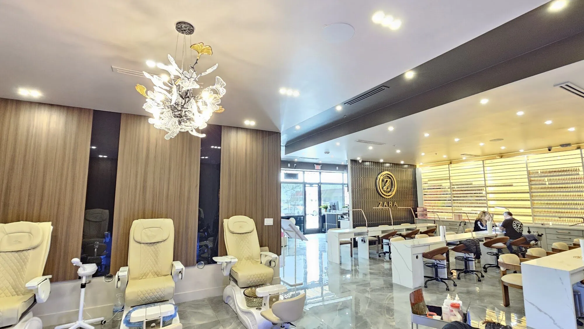 Cream pedicure chairs under a glass chandelier at Zara Nail Bar Vistancia, with the gold Zara sign and a lit wall of colour behind the manicure tables.